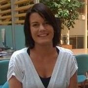 Een vrouw met donker haar glimlacht in een witte top, binnen in de buurt van een plant.