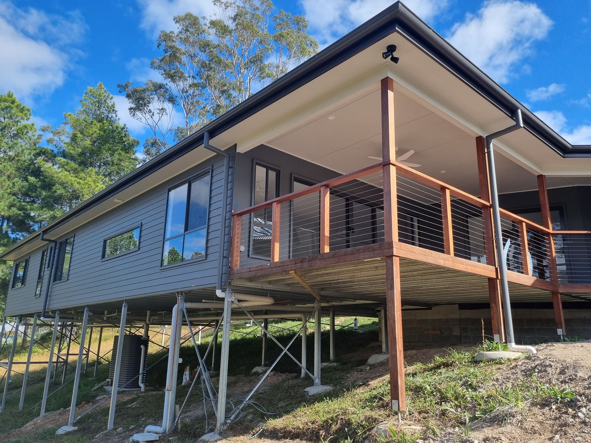 Gray modern house elevated on supports, with a wooden deck and railing. Blue sky, green hillside.