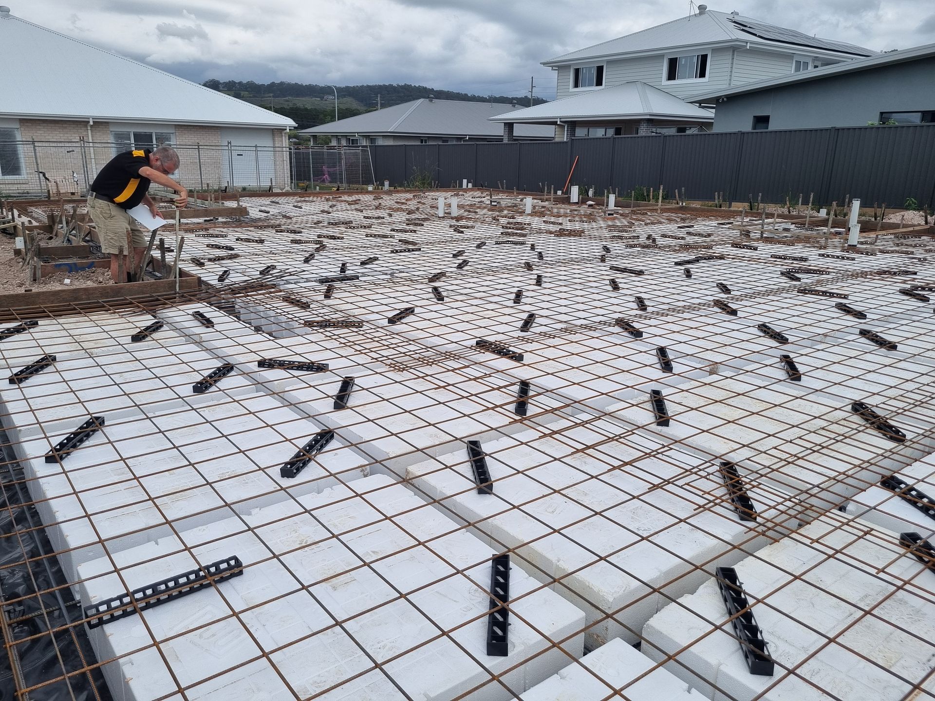Construction site with worker leveling a surface covered in foam blocks and rebar. Cloudy sky, residential area.