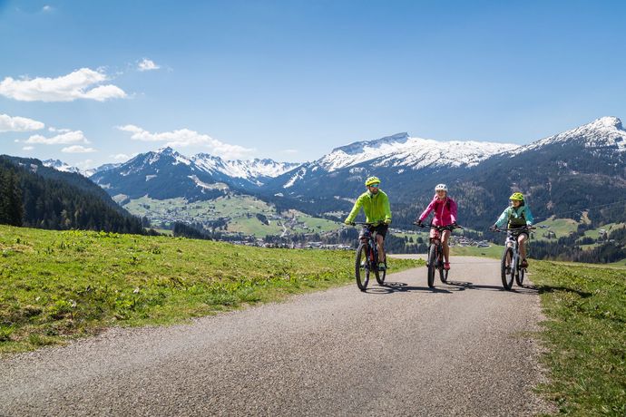 Drei Radfahrer auf einer Bergstraße mit schneebedeckten Gipfeln und blauem Himmel im Hintergrund