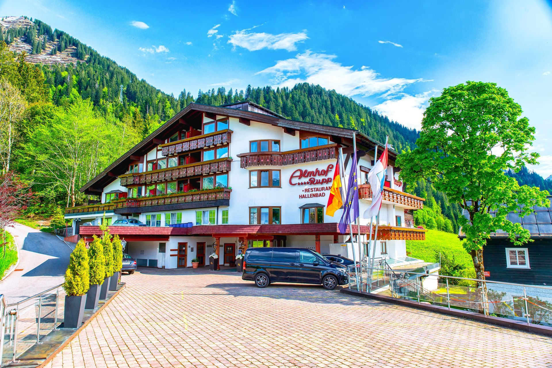 Chalet d'hôtel de montagne avec balcons, parking et collines verdoyantes sous un ciel d'un bleu éclatant