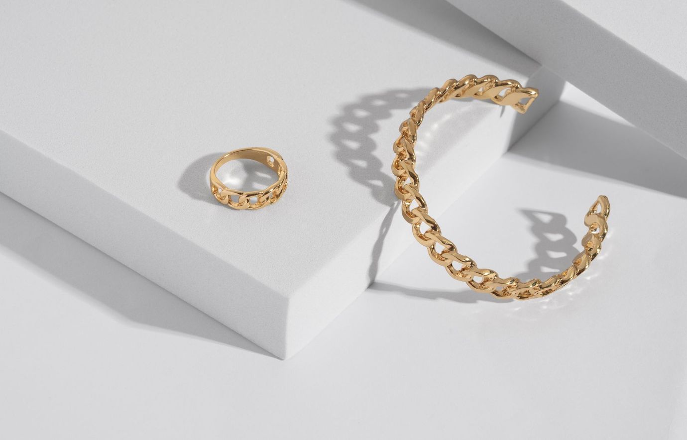 Bracciale e anello a catena in oro su una superficie geometrica bianca.
