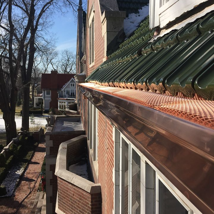 Sunlit brick apartment exterior with green metal roof and narrow walkway beside leafless trees