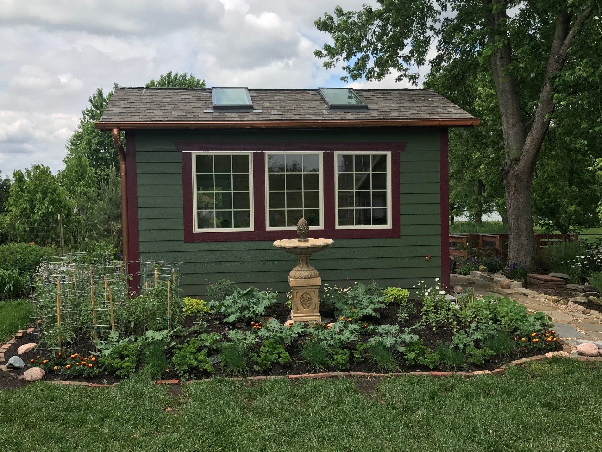 Small green garden shed with red trim, windows, and a birdbath in a flower bed.