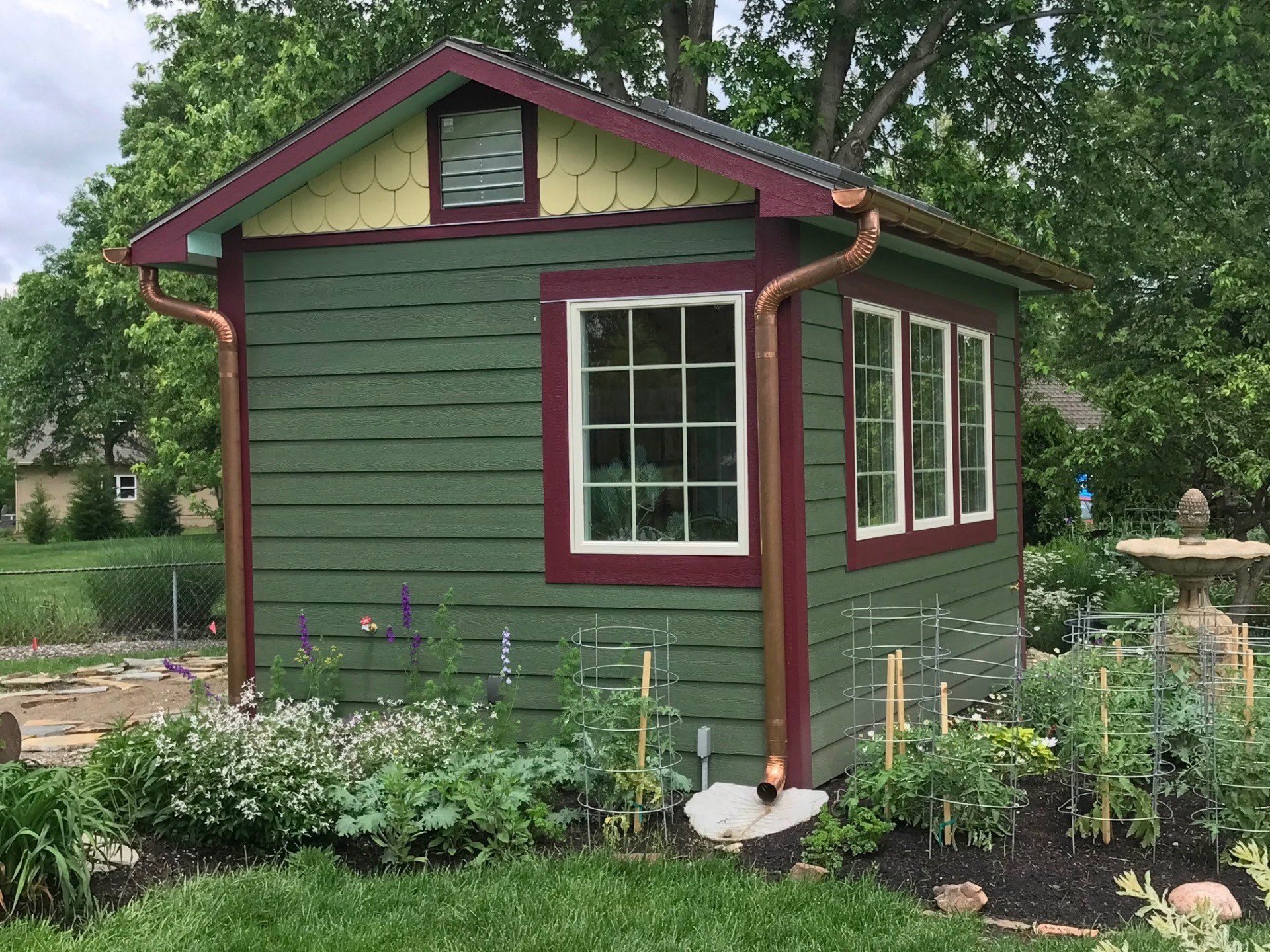 Small green garden shed with maroon trim and white windows, surrounded by flowers and trees.