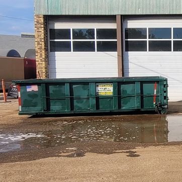 Green dumpster in front of a building with garage doors. Puddle in front of the dumpster.