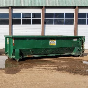 Green dumpster in front of a building with windows and garage doors, sitting on a muddy surface.