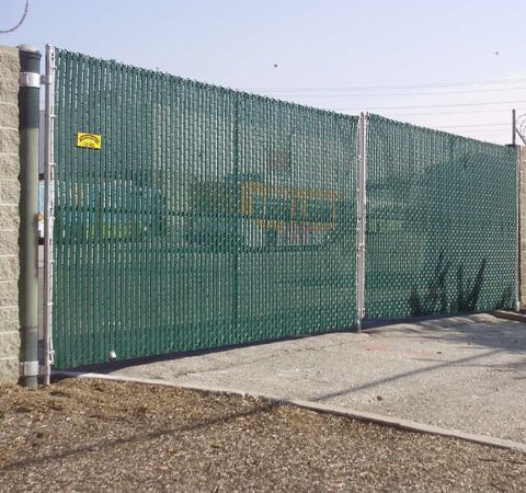 Green chain-link fence along a concrete sidewalk beside a gravel lot under a clear sky