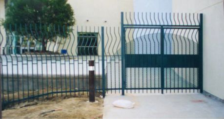 Dark green metal fence and gate in front of a house with a tree and sidewalk.