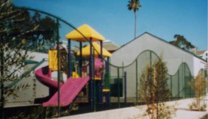 Playground with pink slide and yellow towers beside a white building and fence under a blue sky