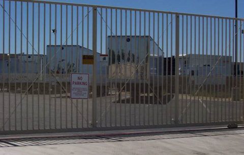 Tall metal gate with a warning sign in front of an industrial building and chain-link fence.