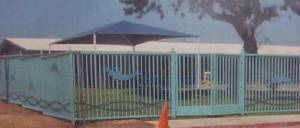 Light blue metal fence around a small building, with a tree and an orange cone in front.