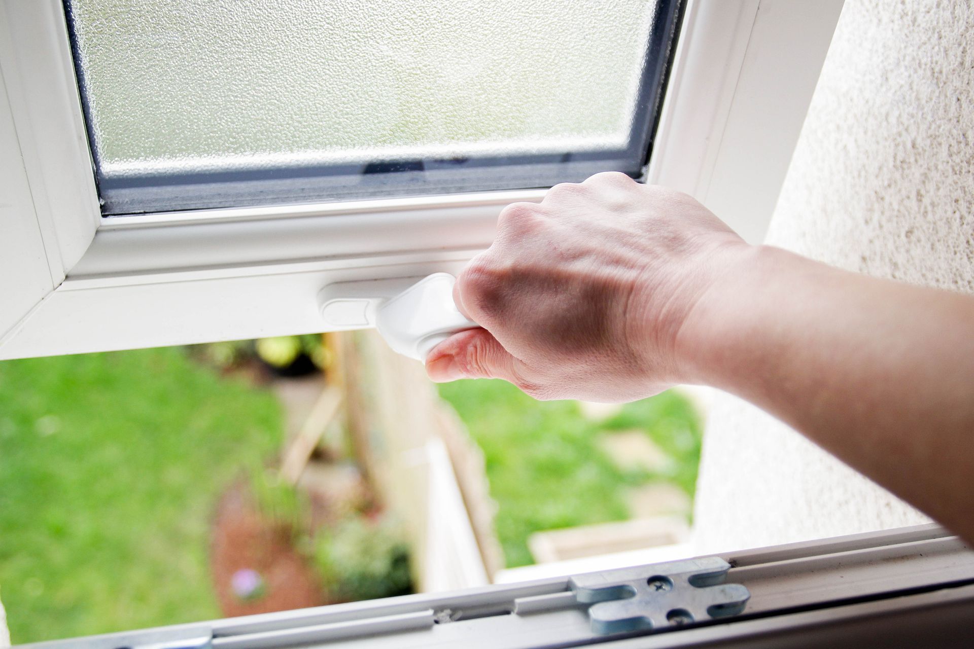 A hand gripping a white handle to open a double-glazed window with frosted glass and a garden view