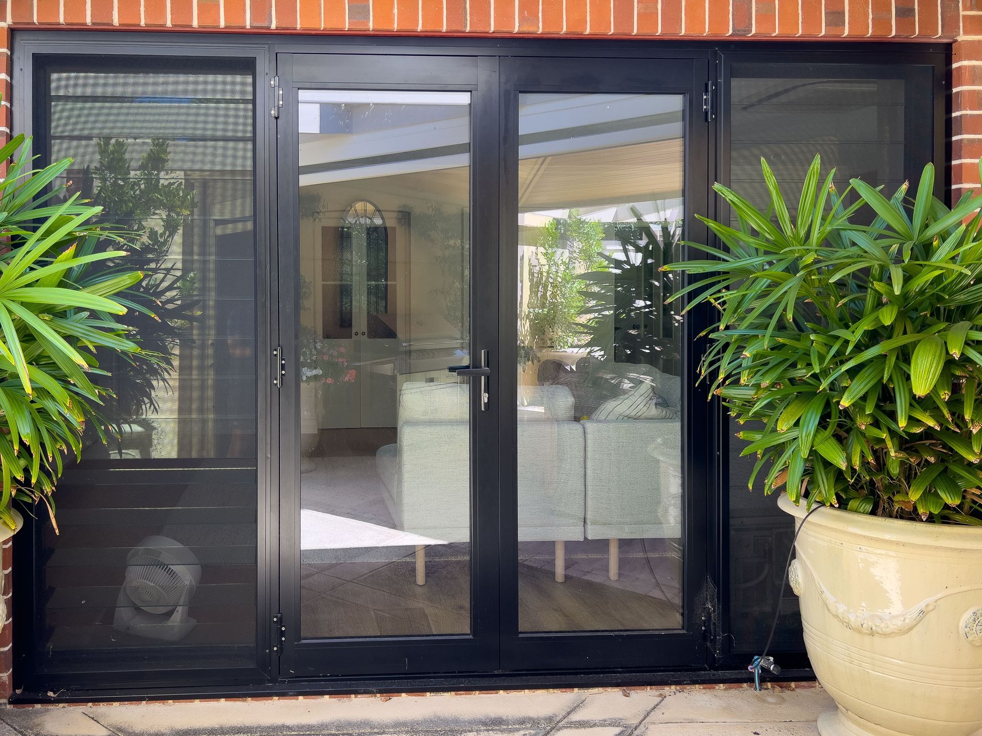 Black French doors with glass panes and screen doors, flanked by potted plants, set against a brick wall.