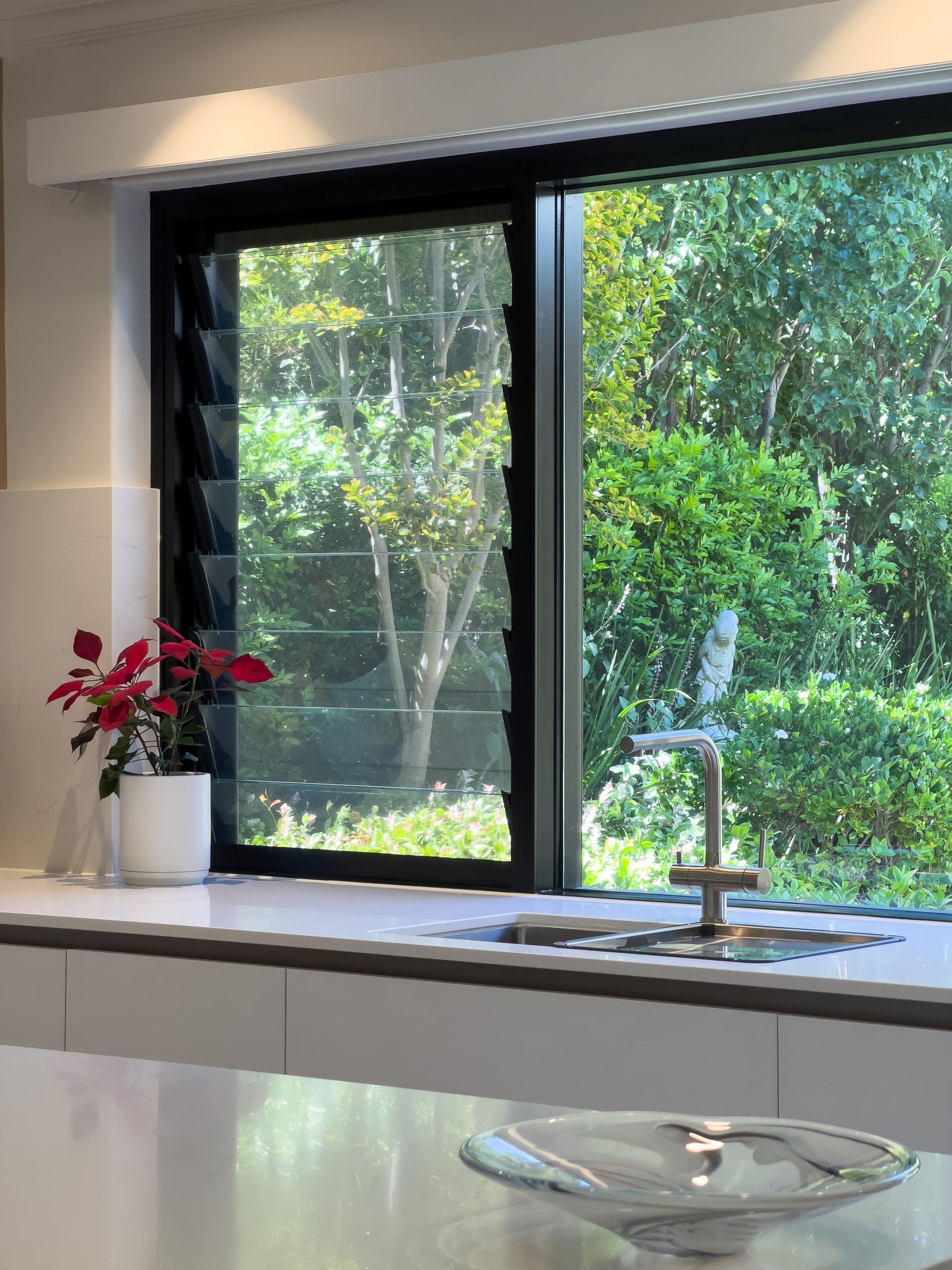 Kitchen window with a view of lush green trees, a sink, and a red potted plant.