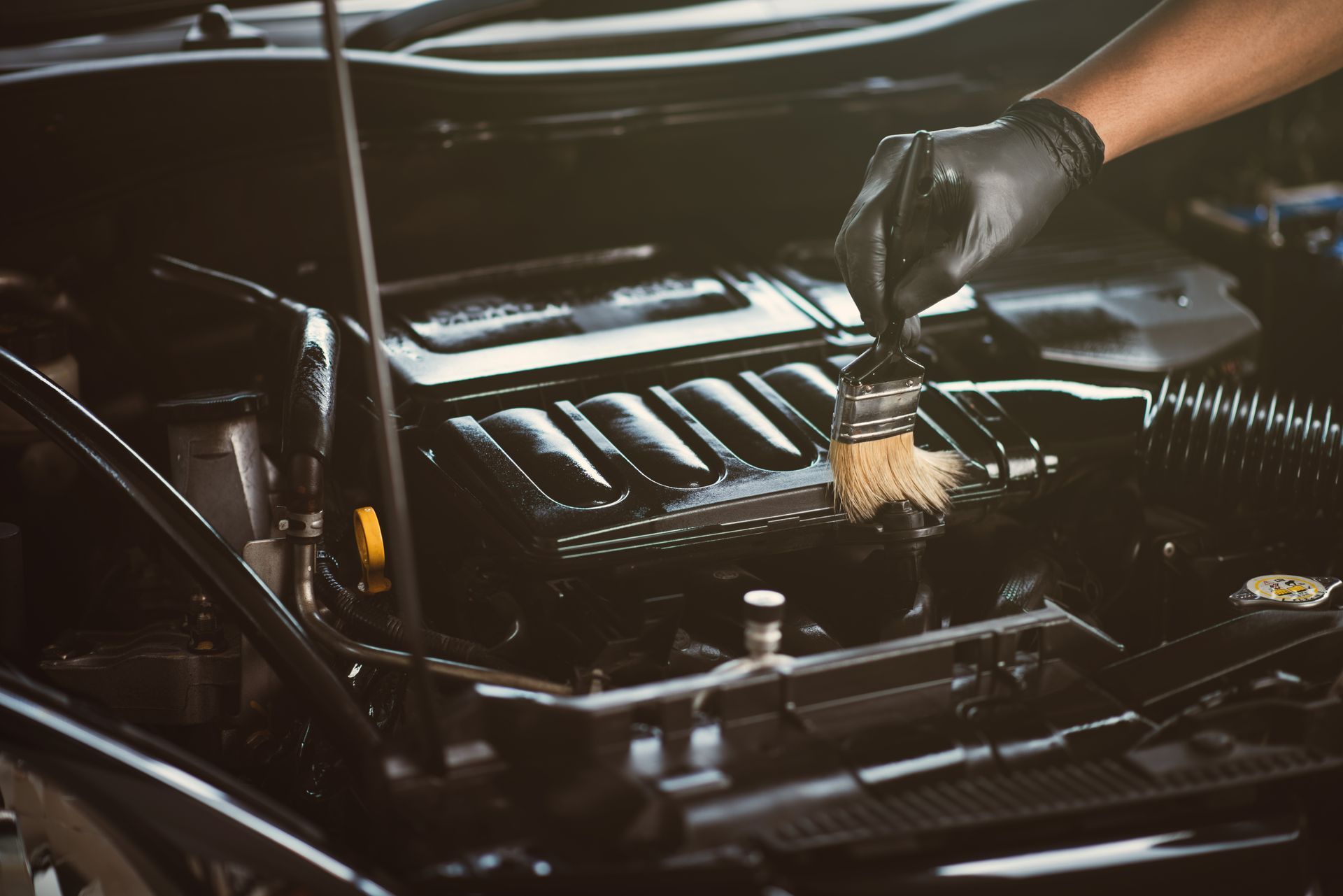 Black gloved hand uses a brush to clean a car engine.