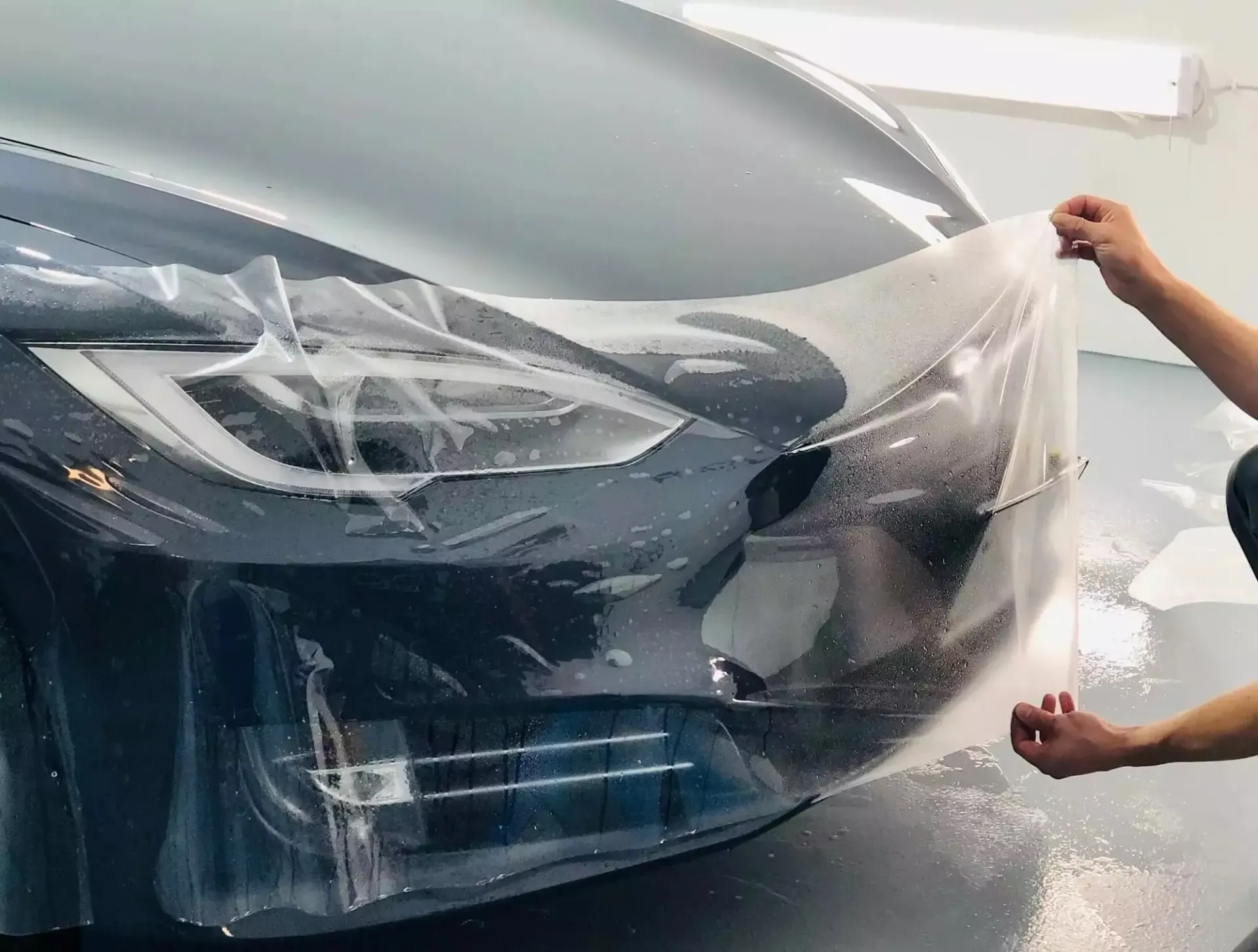 Person installing clear protective film on a dark-colored car's front bumper and headlight.