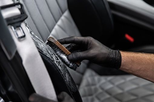 Person wearing black gloves cleaning car seat with brush.