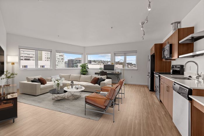A modern, spacious apartment interior with a living area, kitchen, and windows with city views. Furnishings include a large sectional sofa, chairs, and a desk.