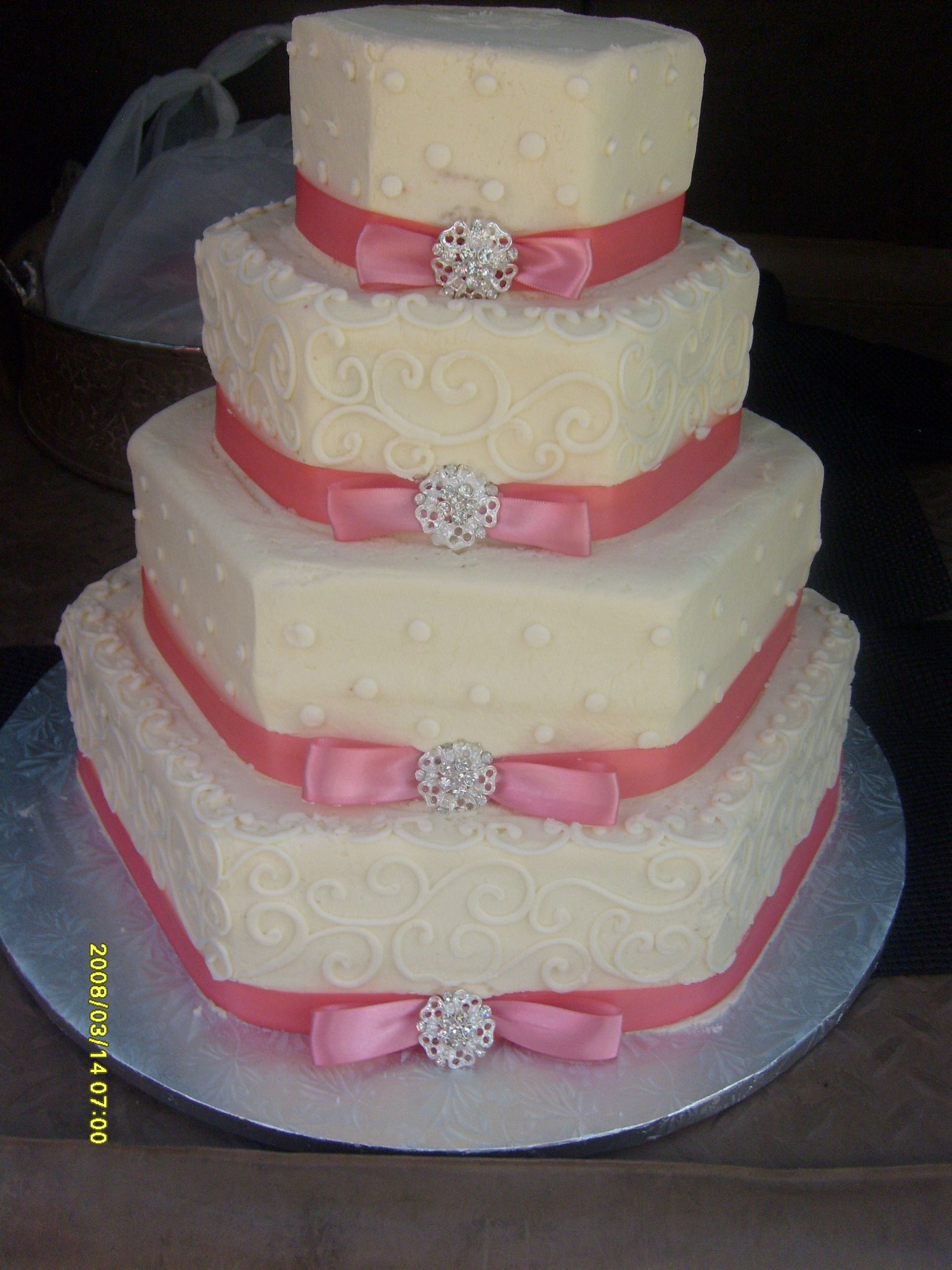 Four-tier ivory wedding cake with pink ribbon bands and silver brooch accents on a silver board