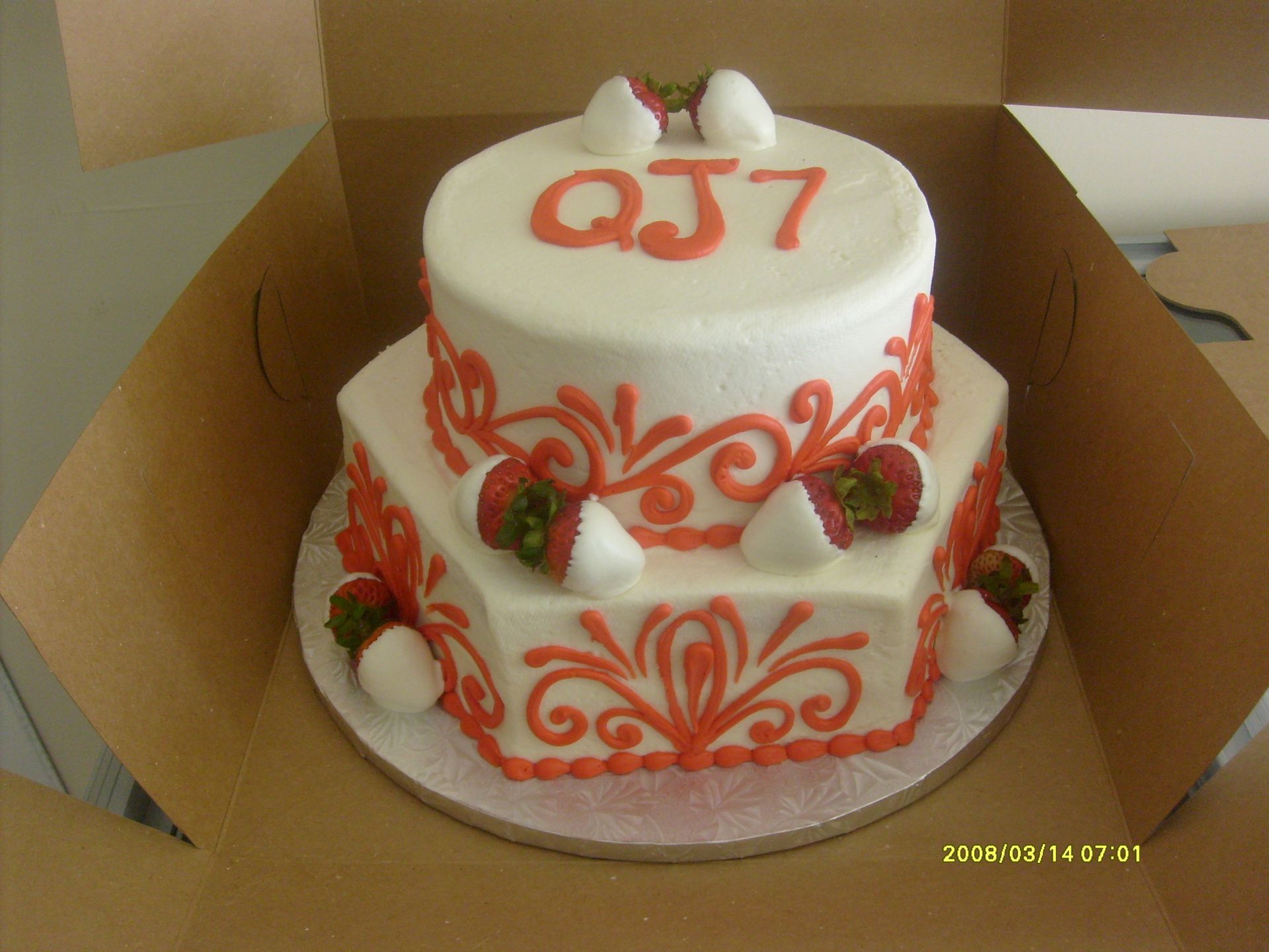 Decorated white cake with red accents and berries, labeled “QJ7,” in a cardboard box