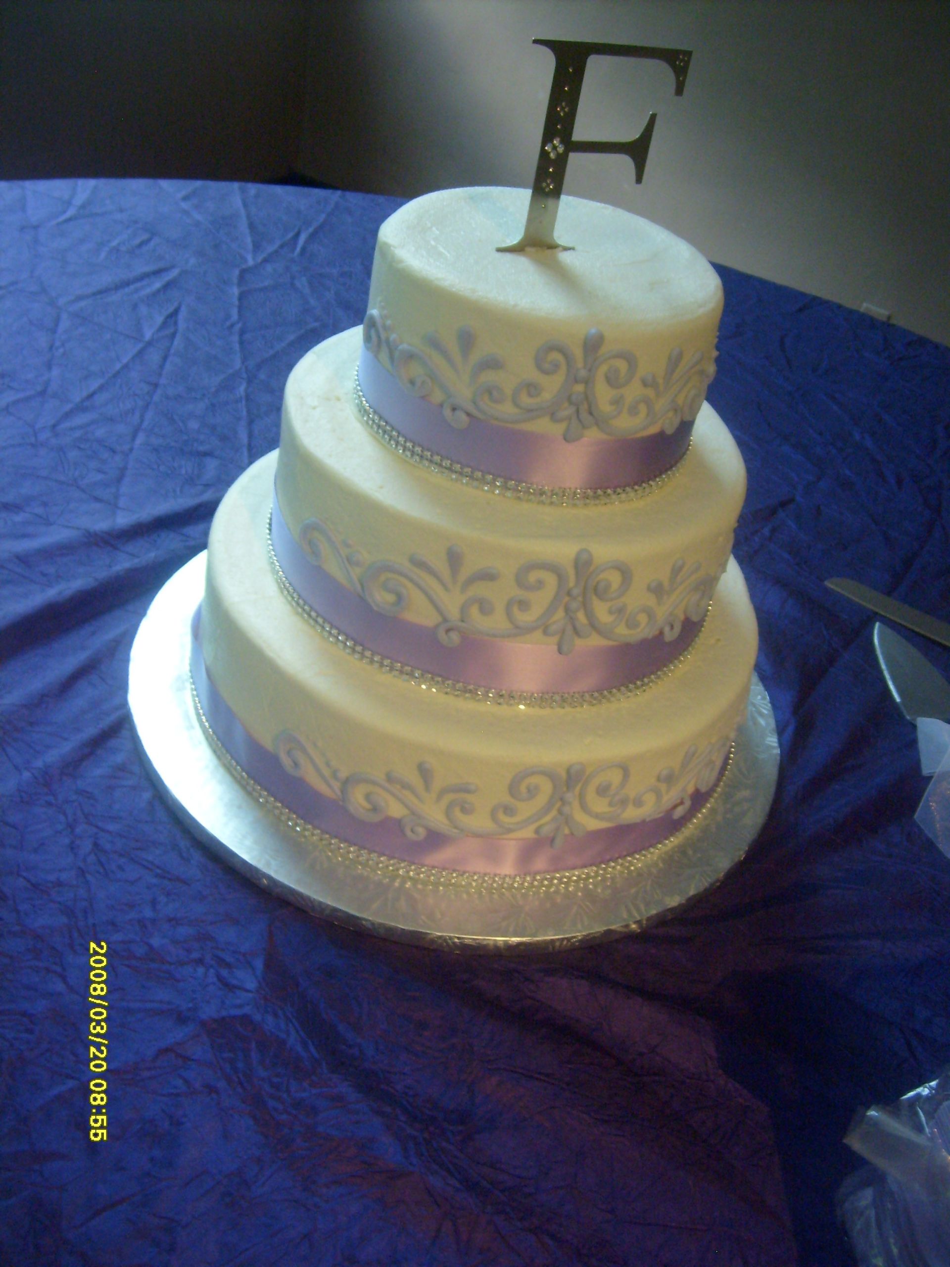 Three-tier white cake with lavender ribbon and a gold letter F topper on a silver board.