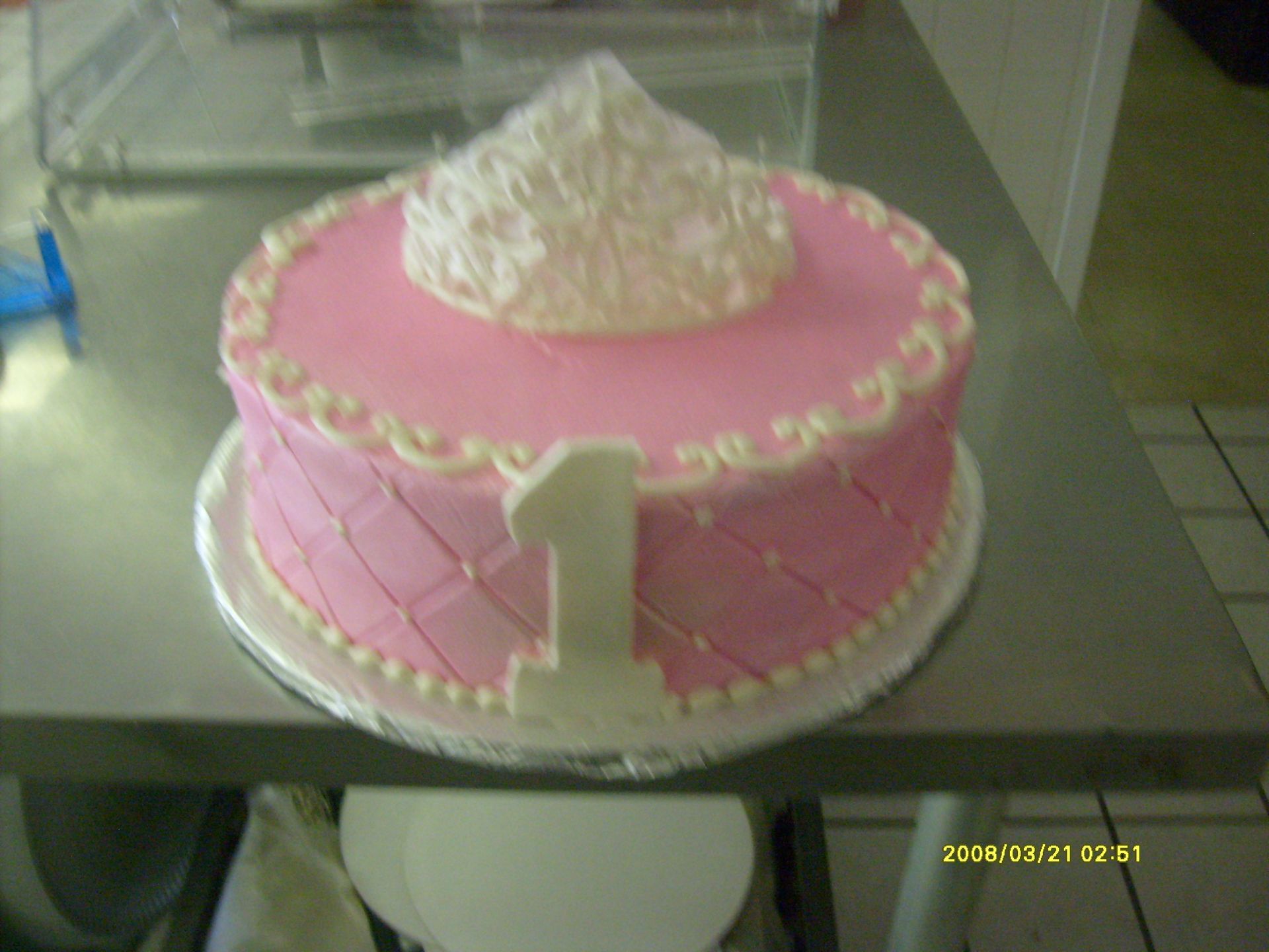 Pink frosted cake with a large white number 1 on top, displayed on a tray behind glass.