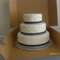 Three-tier white cake with dark ribbon bands in an open cardboard box