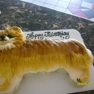 Yellow furry birthday cake shaped like a dog with “Happy Birthday” written on top