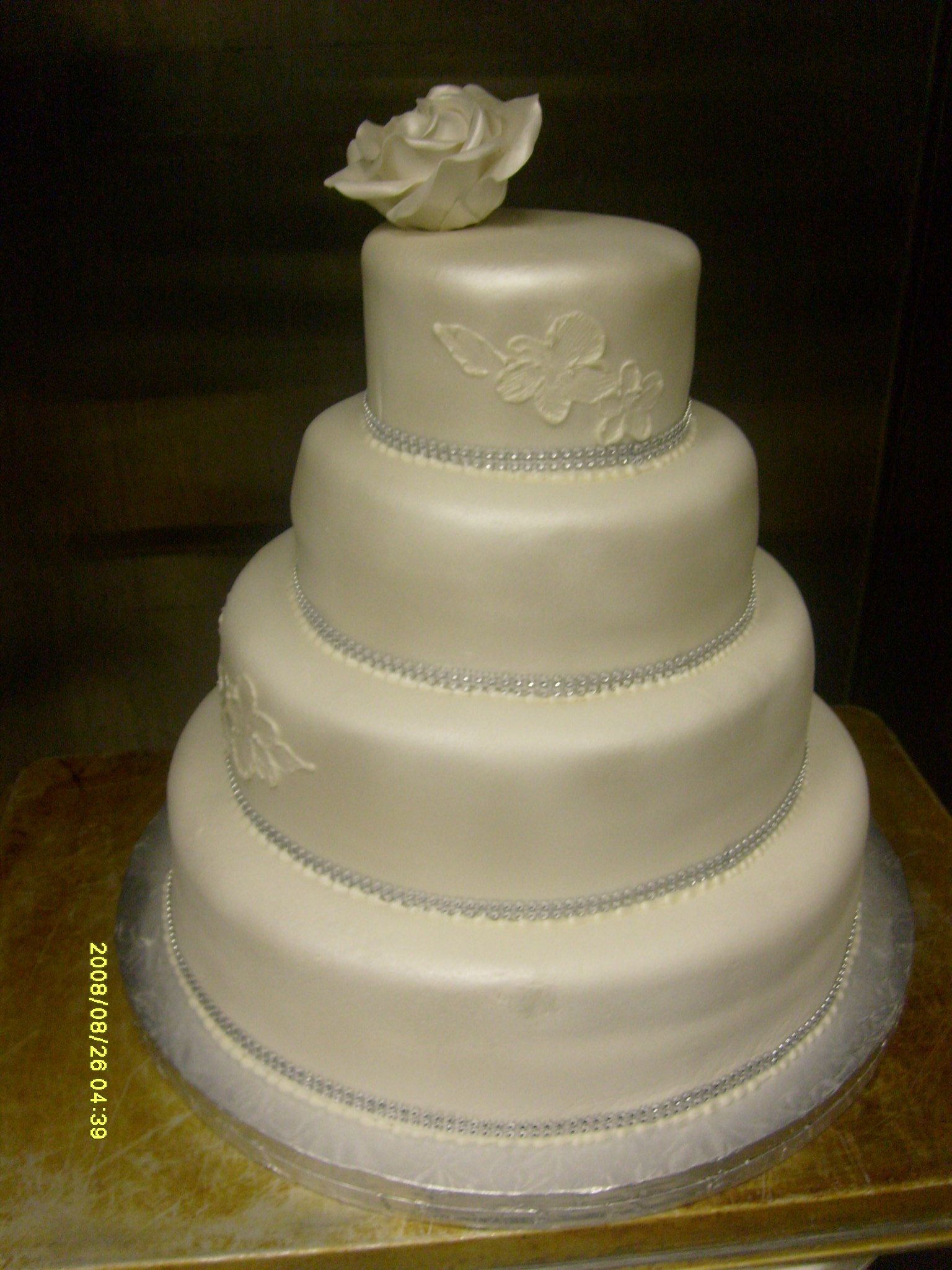 Four-tier white wedding cake with silver trim and a large rose topper