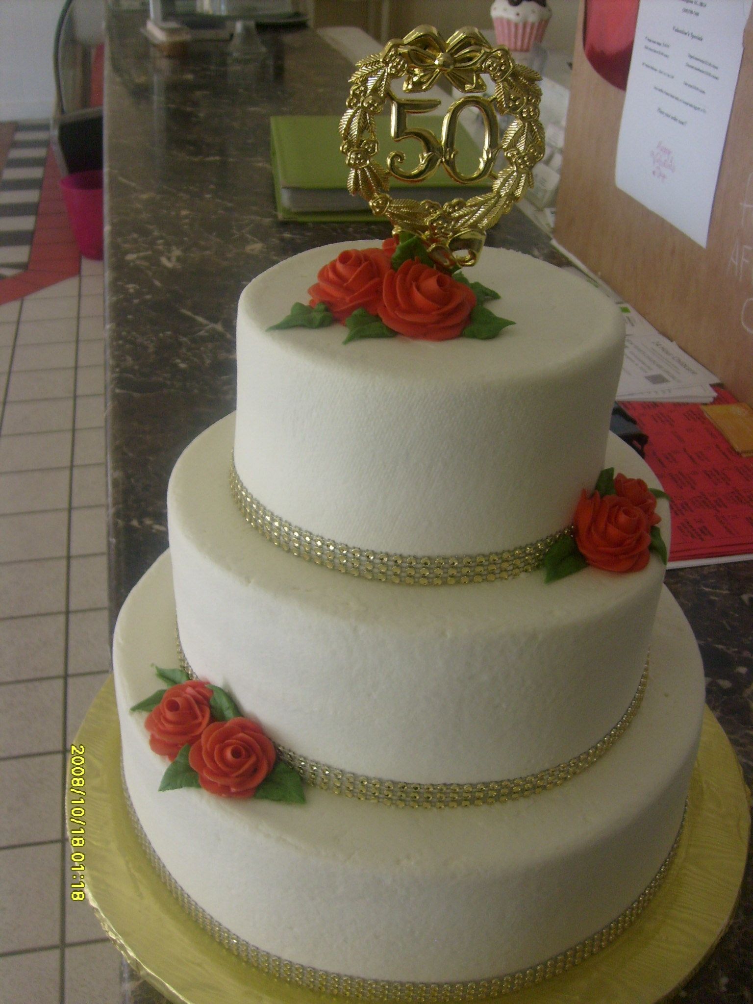 Three-tier white cake with gold accents and red roses, topped with a gold 50 decoration