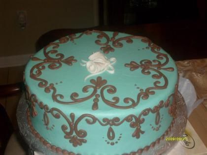 Turquoise frosted cake with brown scroll decorations and white flower topper on a table