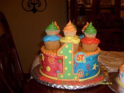 Colorful birthday cake shaped as a large number 1 with cupcakes on top, on a table indoors
