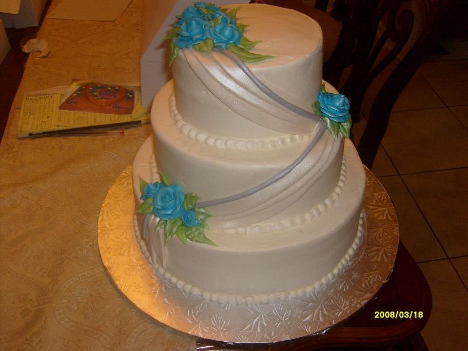 Three-tier white wedding cake with blue flowers and silver ribbon accents on a gold cake board
