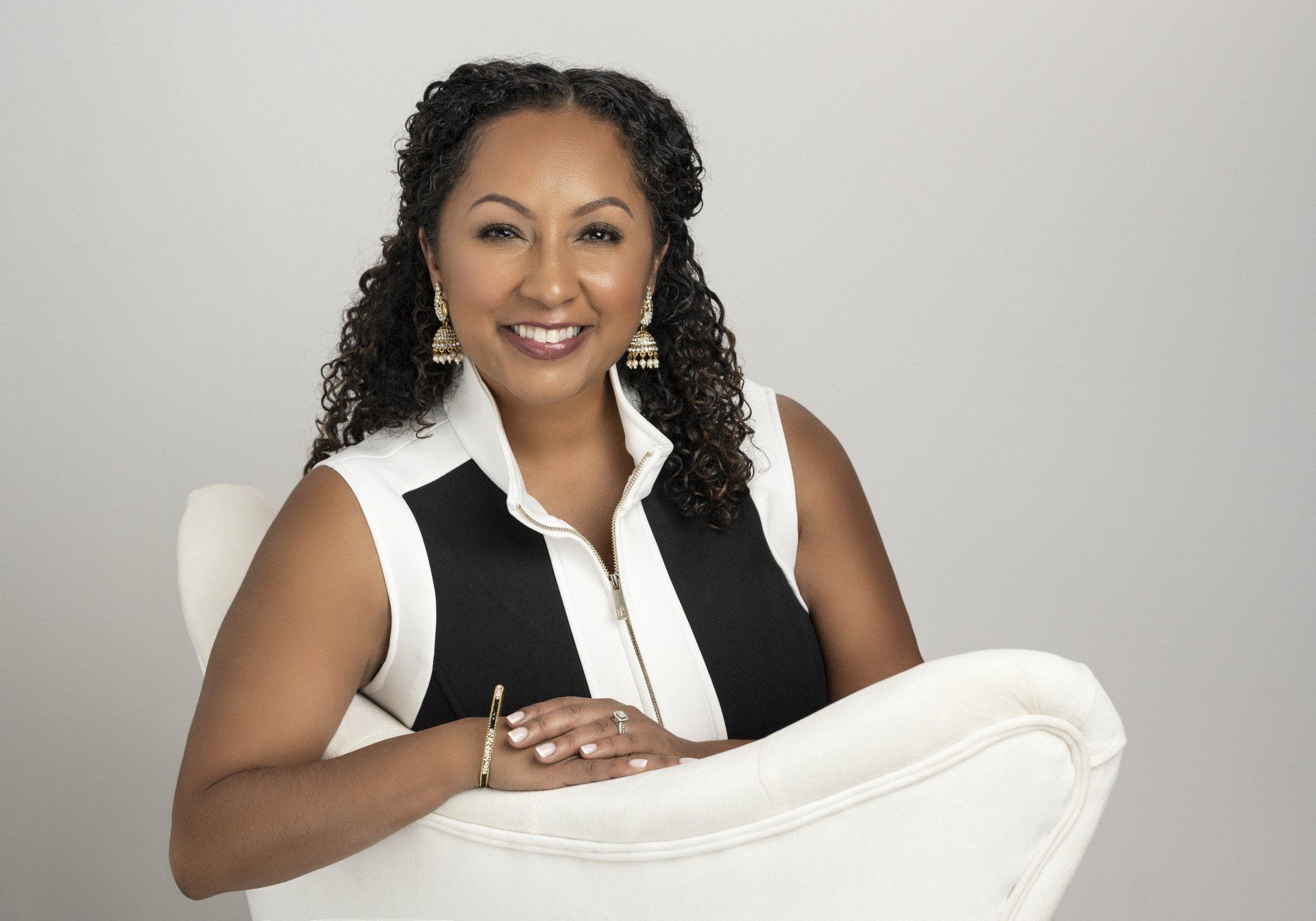 A woman is sitting in a white chair with her hands folded and smiling.