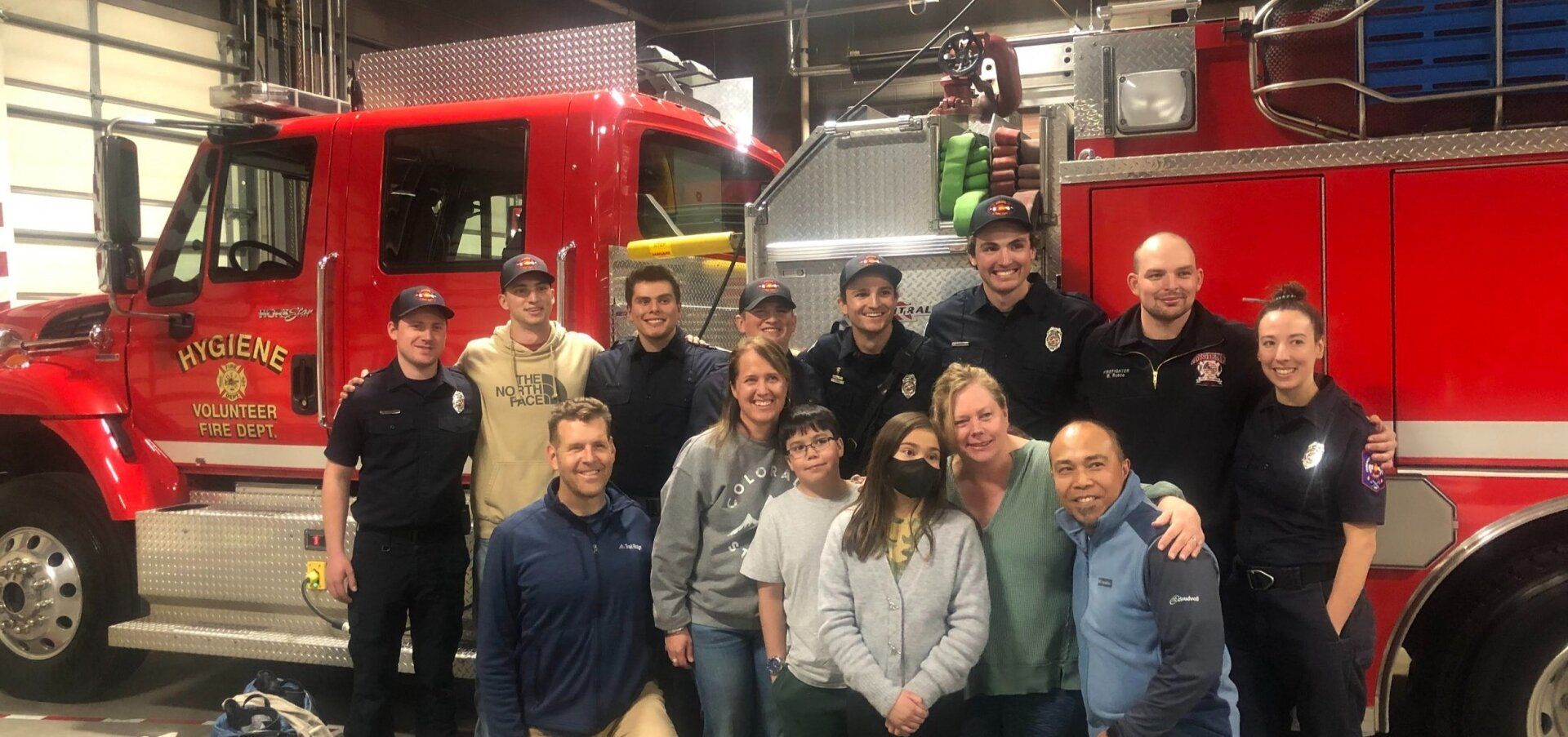 A group of people huddle in front of a fire truck. A mix of firefighters and civilians, they are all smiling