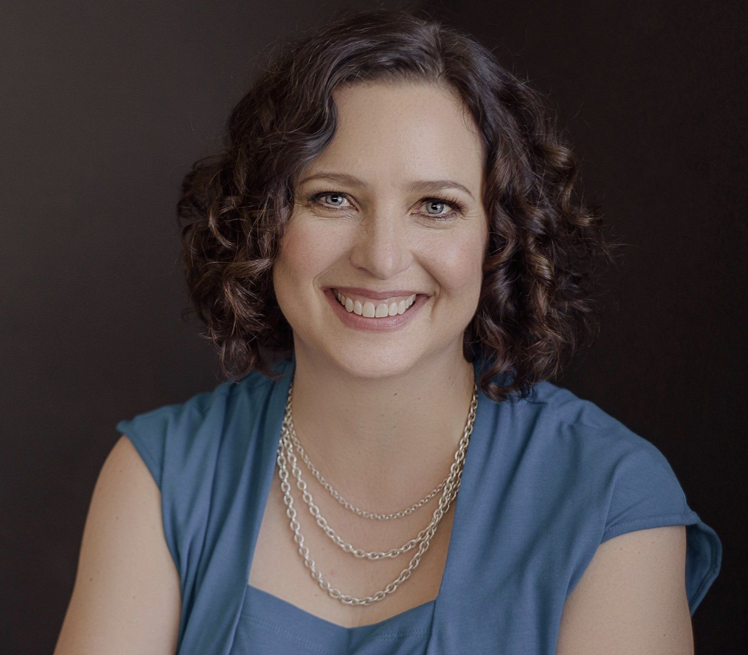 A woman wearing a blue shirt and a necklace is smiling for the camera.