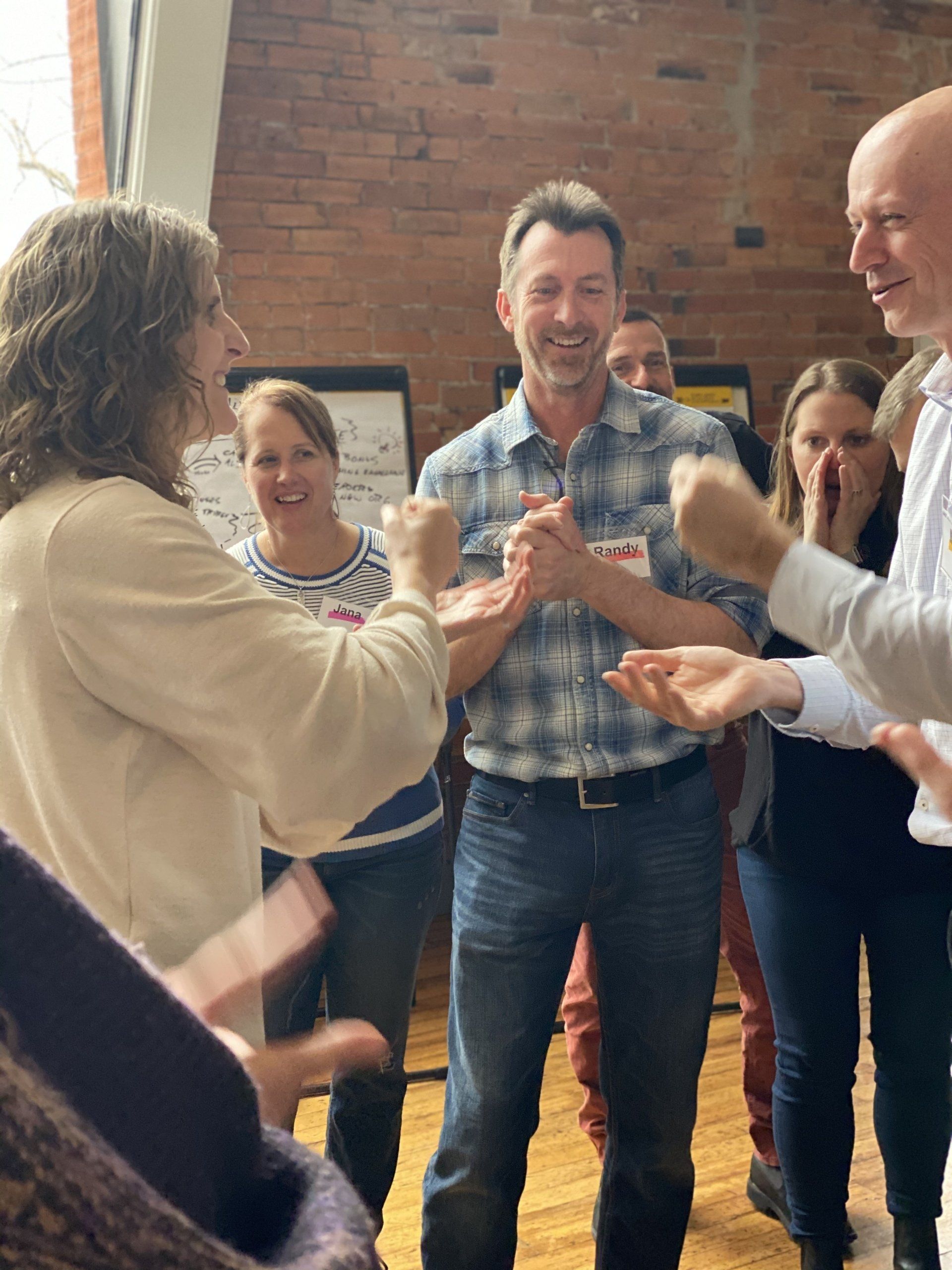 A group of men and women gather in a circle. Two are playing rock, paper scissors while the others cheer them on.