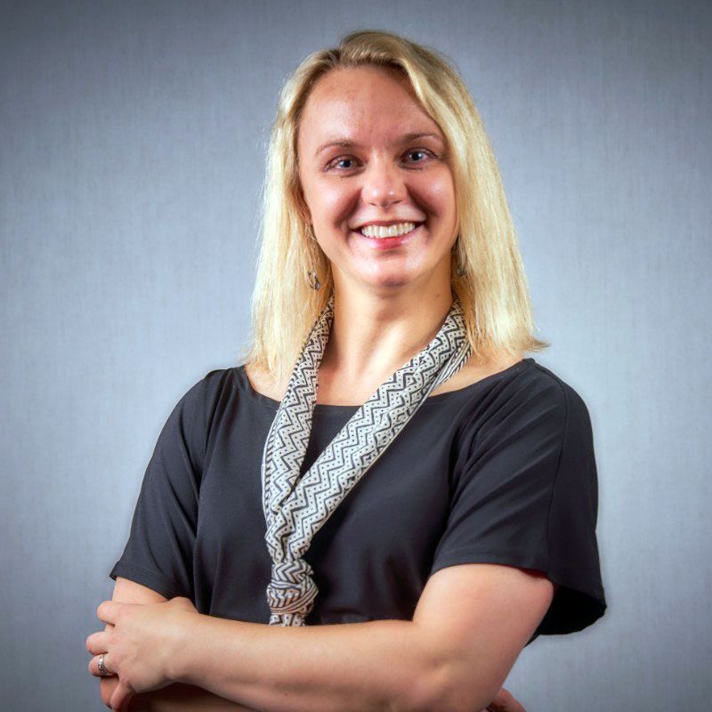 Katharine Bodan smiles, arms crossed; studio portrait against a gray background.