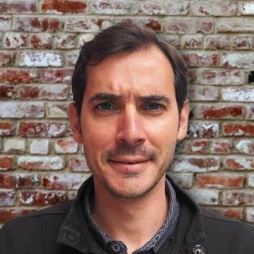 A man with a beard is smiling in front of a brick wall.