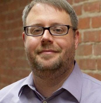 Man with glasses and a beard smiles, set against a brick wall, wearing a lavender shirt.