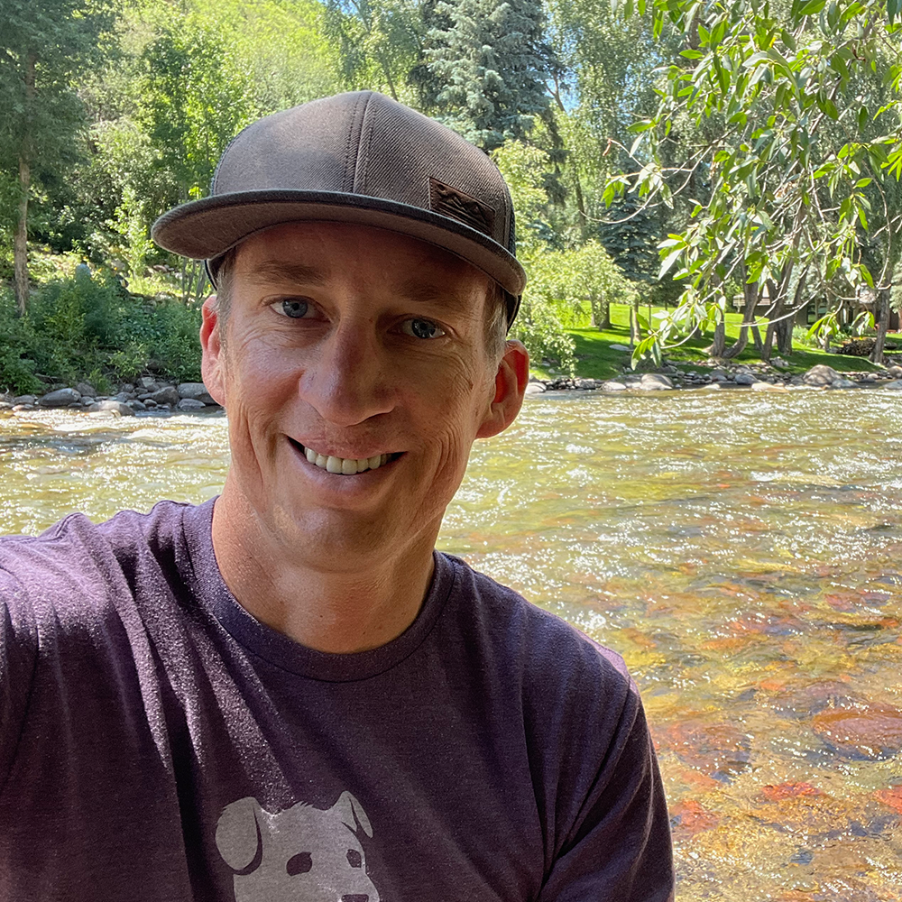 A man wearing a hat and a purple shirt is smiling in front of a river.
