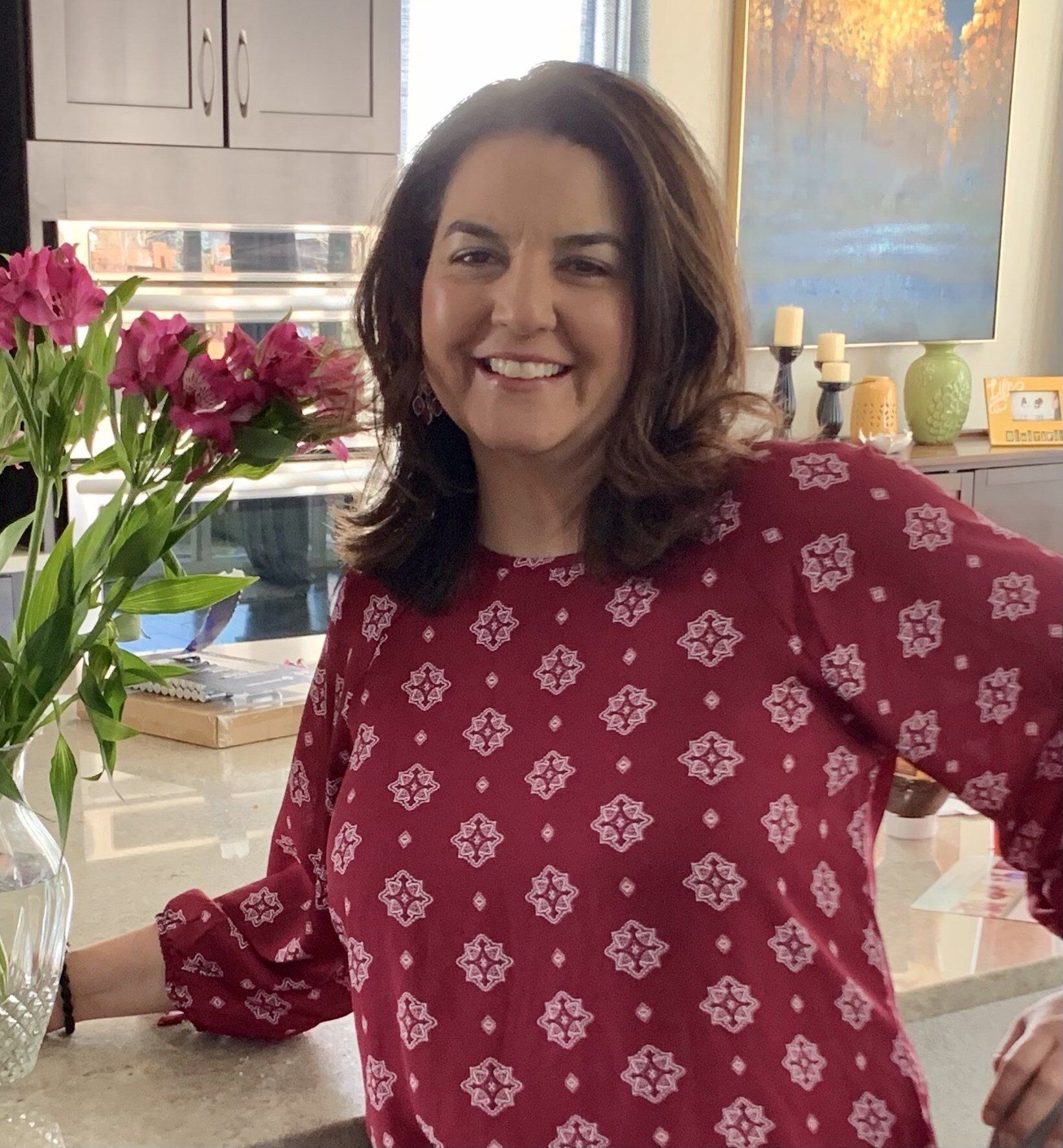 A woman in a red shirt is smiling in front of a vase of flowers