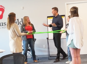 Four people hold a green rubber band in a room, with a man gesturing. A whiteboard and logo are in the background.
