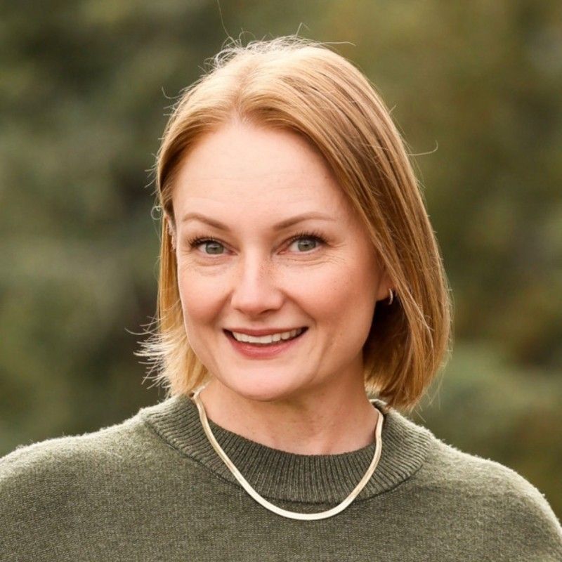 Woman with red hair smiling, wearing a green sweater and necklace, set outdoors.