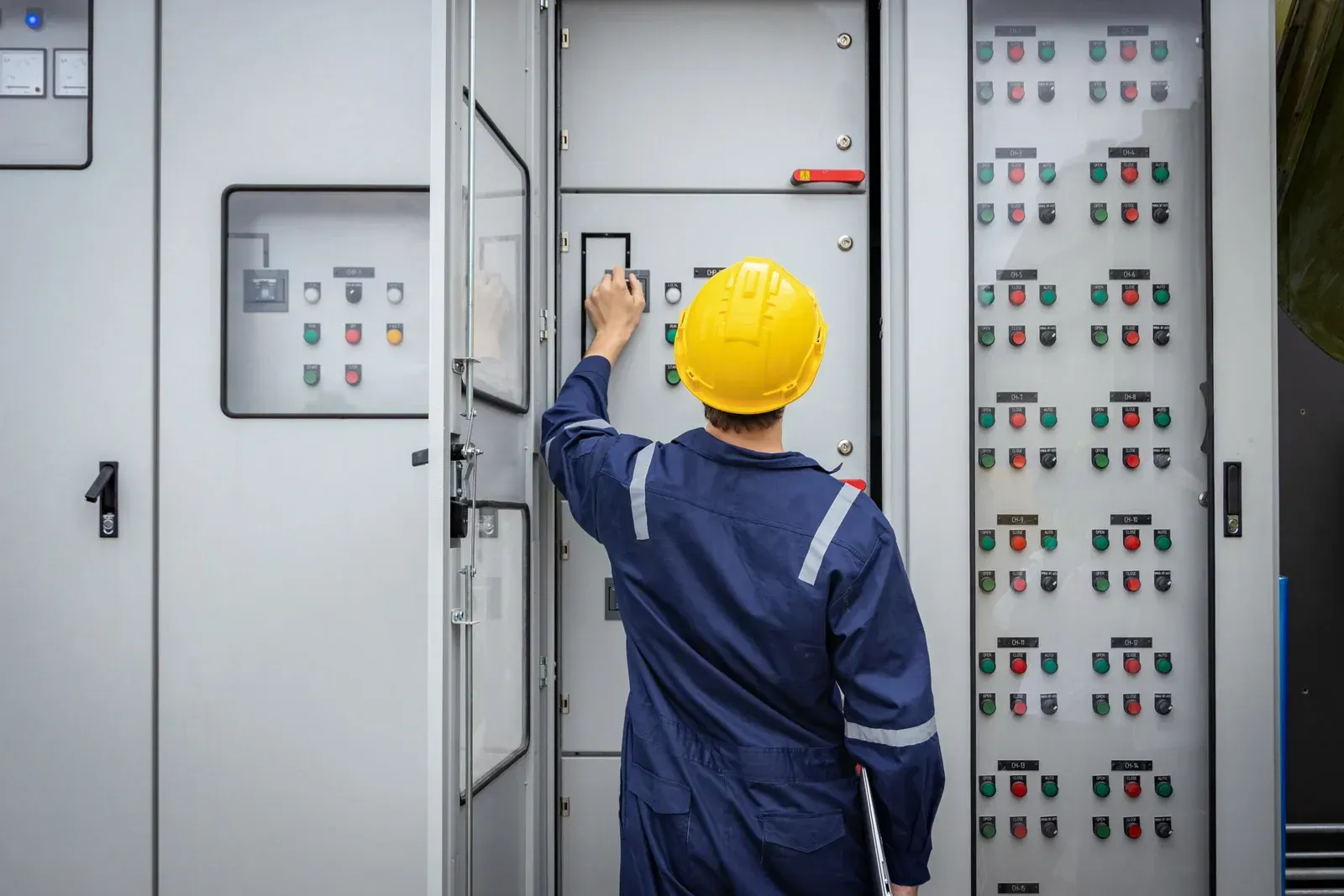 A technician in a blue jumpsuit and yellow hard hat opens an electrical panel, inspecting the components.