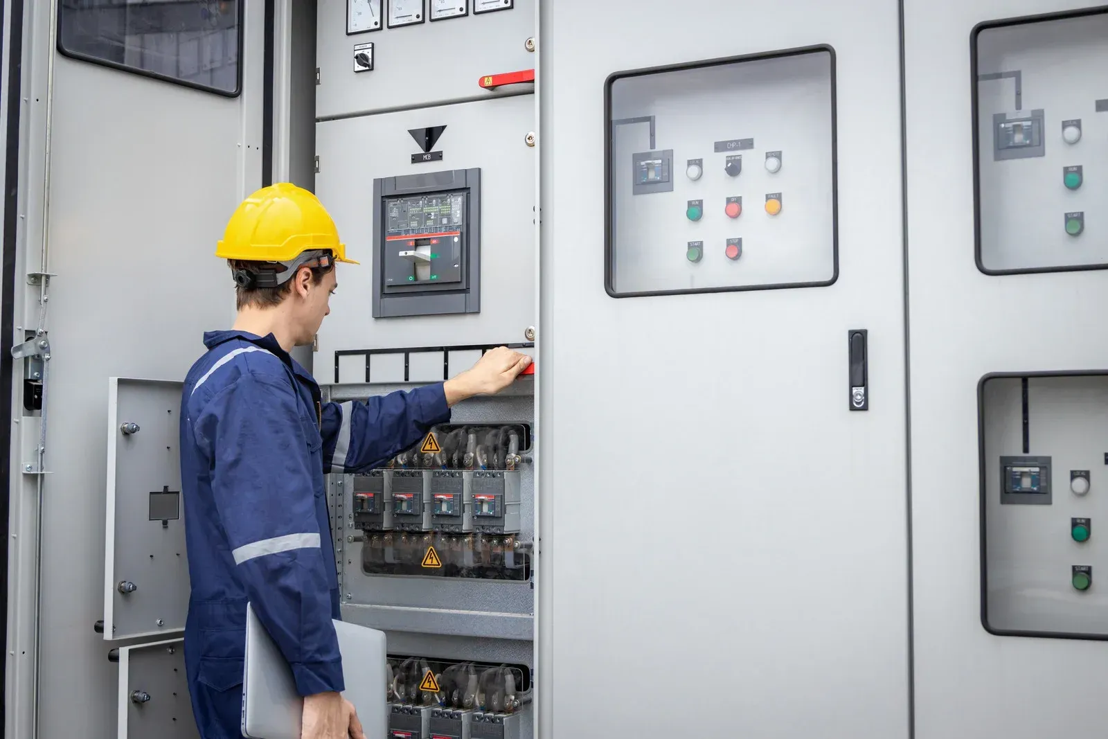 Electrician in a yellow hard hat and blue jumpsuit working on electrical panel.