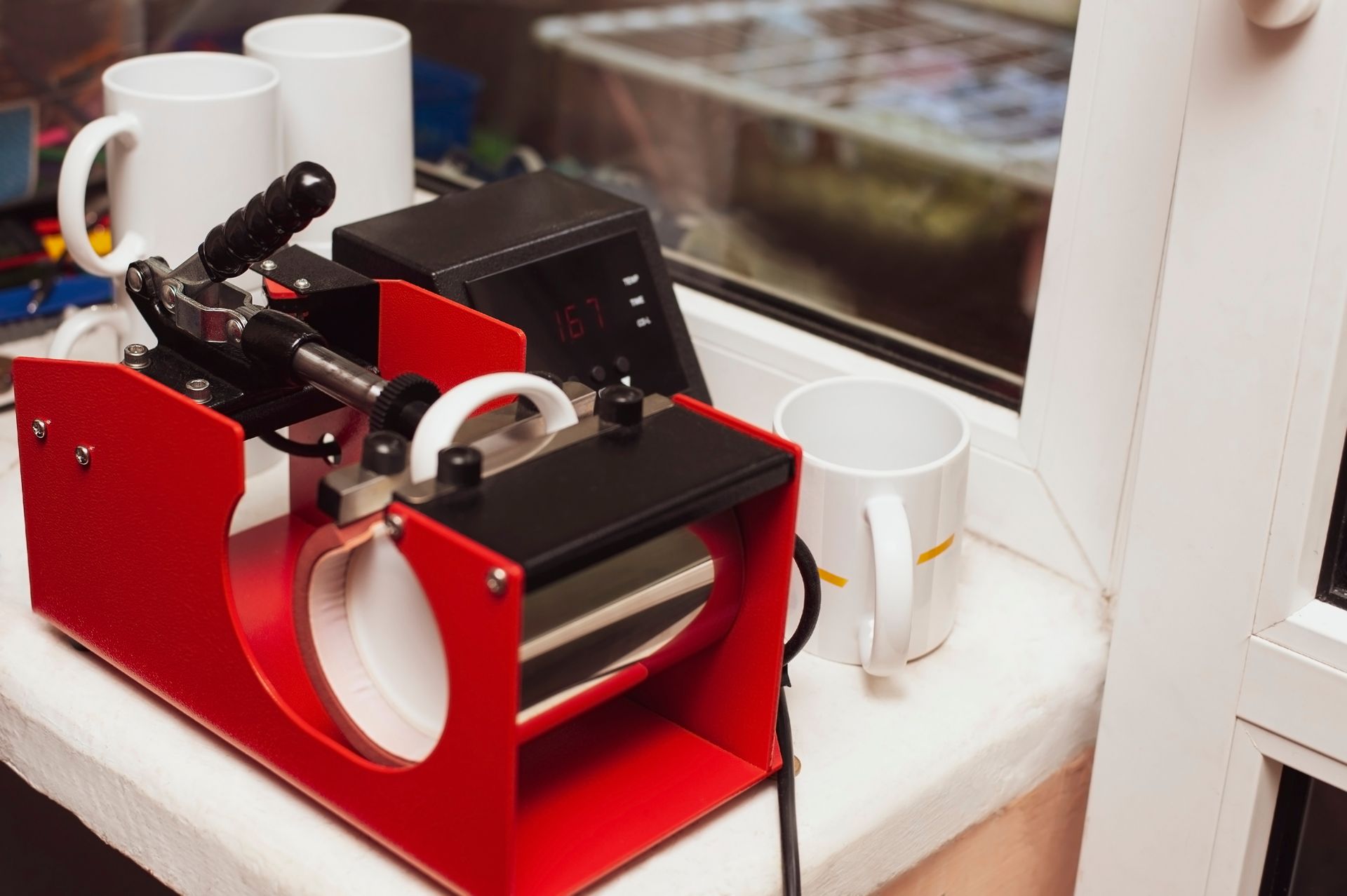 Red mug heat press machine on a windowsill with white ceramic mugs placed nearby for printing.