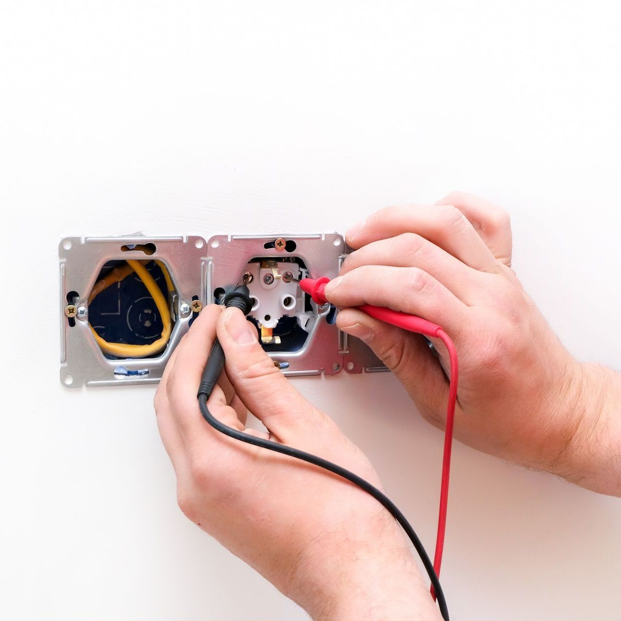 Hands testing an electrical outlet with a multimeter, against a white wall.