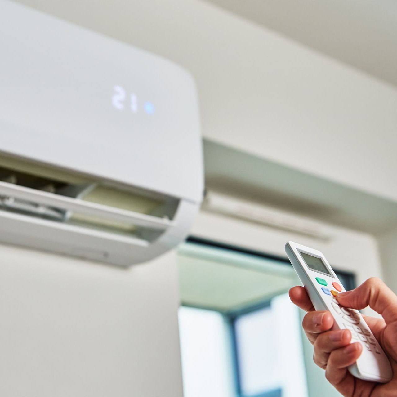Hand holding remote, pointing at wall-mounted air conditioner displaying 21 degrees.